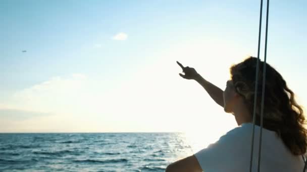 Молодая кудрявая девушка на парусной яхте показывает палец на самолете, взлетающем на закате — стоковое видео