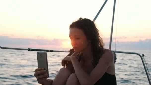 Jonge vrouw is zeiljacht en maakt gebruik van een smartphone - werpt een selfie bij zonsondergang — Stockvideo