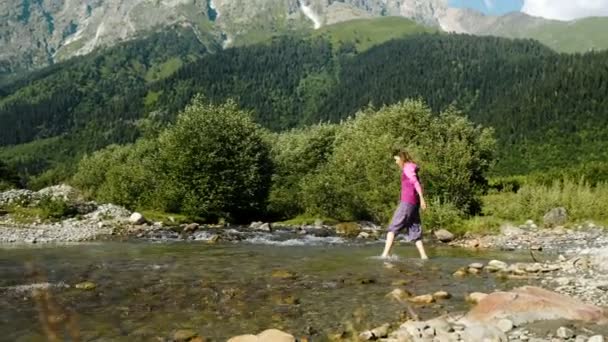 Mulher turista atravessa o riacho no fundo de uma paisagem de montanha — Vídeo de Stock