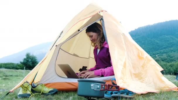 Glad turist flicka använder en bärbar dator i ett tält, frilansare som skriver ut i naturen på en resa — Stockvideo