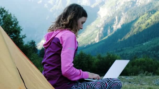 Mulher turística usa um laptop na natureza, freelancer digitando em um computador perto de uma tenda em uma viagem contra o pano de fundo de uma paisagem montesa close-up — Vídeo de Stock
