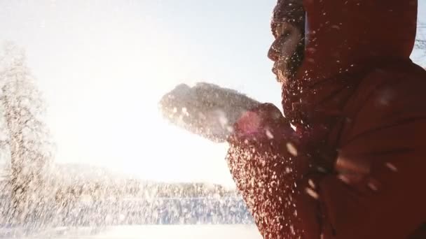 Linda chica en una chaqueta roja soplando copos de nieve de sus palmas en los mitones en invierno contra el atardecer, cámara lenta — Vídeo de stock