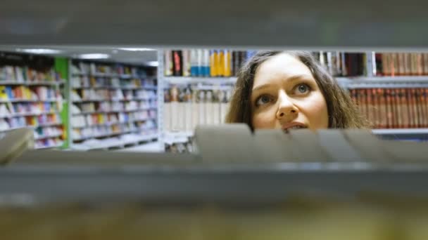 Mooi meisje kiest een boek in de boekhandel, beweegt de camera door middel van de boekenplank — Stockvideo