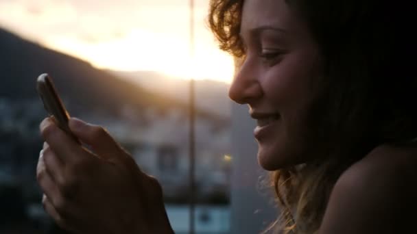 Mujer sonriente con el pelo rizado utiliza el teléfono en una puesta de sol, sostiene en las manos en la ciudad — Vídeos de Stock