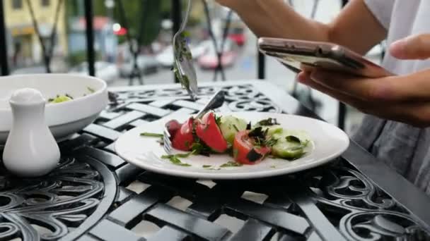 Vrouw zet plantaardige salade in een bord en maakt gebruik van de telefoon tijdens het diner in een restaurant — Stockvideo