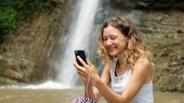 Kadın telefonunu dalgalar, kulaklık video bağlantısı üzerinden görüşmeler ve bir şelale gösterir