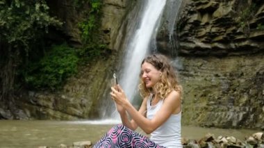 Kadın kulaklık arka planda büyük bir şelale, doğada bir smartphone cep telefonu ile video seslenmek yolu ile iletişim kurmak için kullandığı bir güzel görünüm gösterir