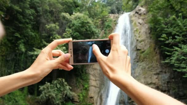 Vrouw richt zich op het scherm van de telefoon en neemt een foto van de waterval close-up — Stockvideo