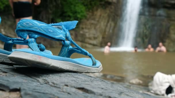 Sandals stand against the background of men bathing in a pond under a waterfall at a defocus — Stock Video