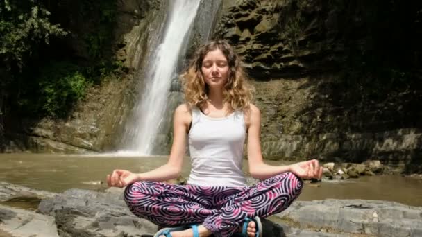 Chica meditando en la posición de loto con los ojos cerrados con una gran cascada sobre un fondo — Vídeos de Stock