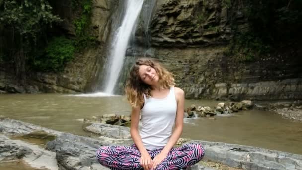 Woman is sitting in the lotus position and doing exercises on the background of a large waterfall — Stock Video