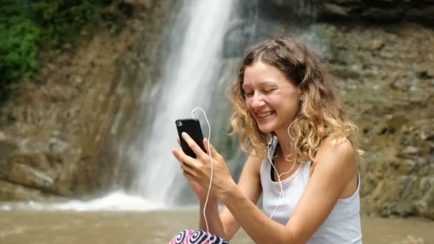 Femme agite son téléphone, parle dans les écouteurs via liaison vidéo, et montre une cascade — Video