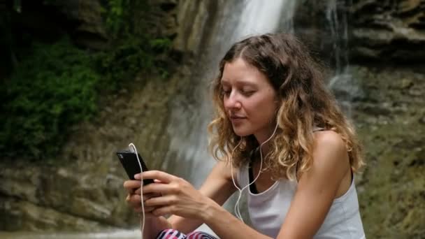 Žena poslouchá hudbu se sluchátky a používá smartphone proti velký vodopád — Stock video