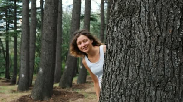 Una chica mira desde detrás de un árbol y le hace señas con un dedo . — Vídeos de Stock