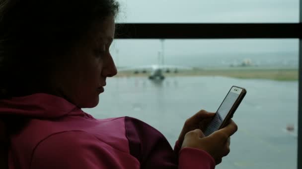 Frau nutzt Smartphone am Flughafenfenster. Flugzeug im Hintergrund — Stockvideo