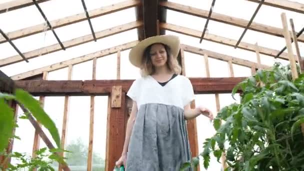 Lachende vrouw boer in een strooien hoed en kleding gaat naar de serre met een gieter — Stockvideo