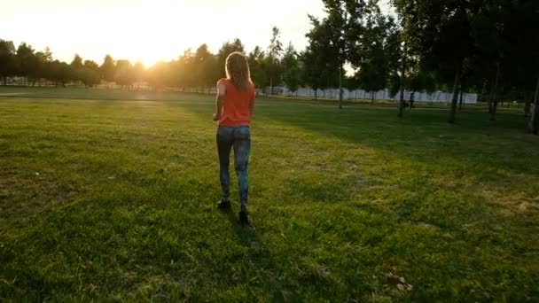 Спортивная девушка бегает по траве на закате в городском парке, замедленная съемка — стоковое видео