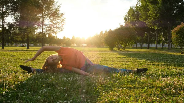 Woman gymnast sits on a string on the grass in a city park in nature and does stretching