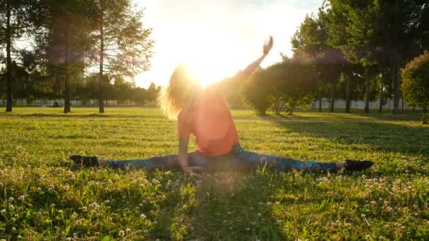 Chica gimnasta se sienta en una cuerda en la hierba en un parque de la ciudad en la naturaleza y hace curvas laterales a las piernas . — Vídeos de Stock