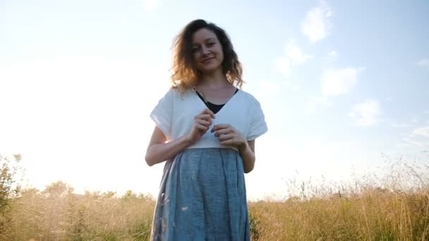 Mulher em um vestido com uma grama na mão caminha em um campo ao pôr do sol, câmera lenta e movimento — Vídeo de Stock