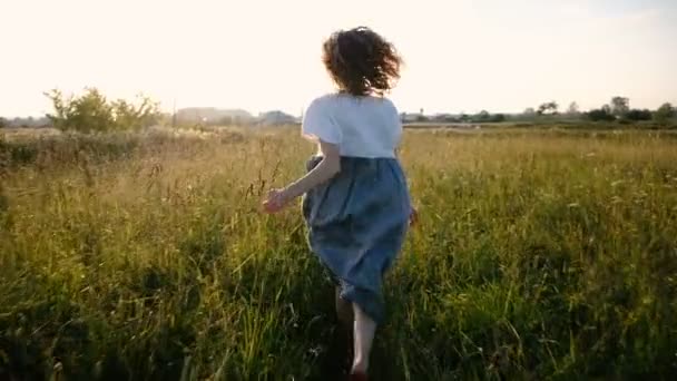 Женщина в платье романтически бежит по полю с высокой травой на закате, замедленной съемкой и движением камеры — стоковое видео
