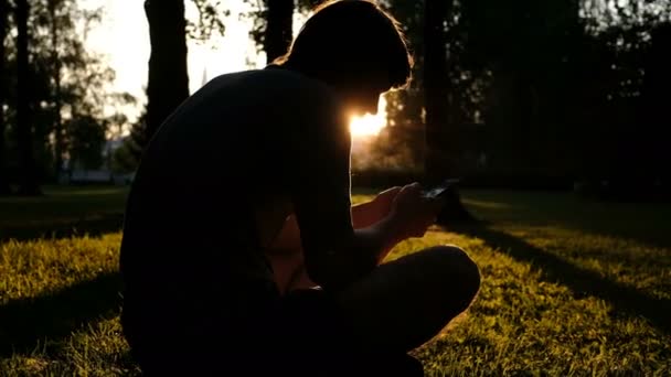 Silhueta de um homem sentado na grama com um smartphone em suas mãos ao pôr do sol no parque — Vídeo de Stock