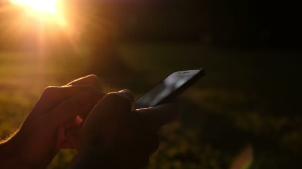 Un uomo tiene uno smartphone tra le mani primo piano al tramonto, i raggi dorati dei soli brillano nel parco . — Video Stock