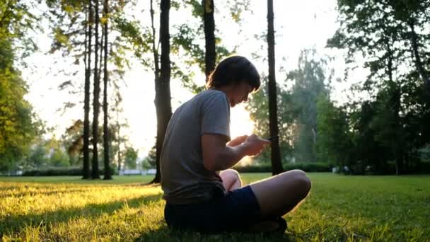 Egy ember használja a smartphone, miközben ül a fűben, egy parkban a naplemente. — Stock videók