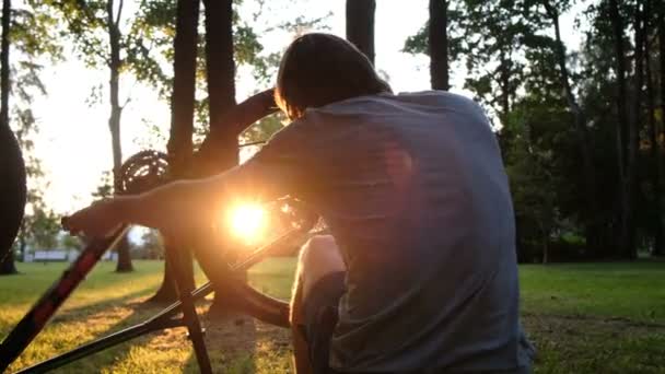 Homem gira e repara uma roda em uma bicicleta virada para cima, o cara repara uma bicicleta ao pôr do sol no parque — Vídeo de Stock