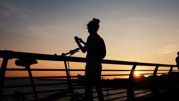 Silhouette of a girl takes pictures of the sunset on the bridge in the summer to the sea — Stock Video