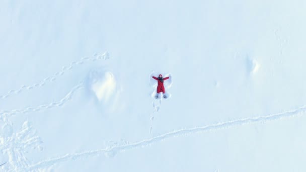 Fotocamera aerea si alza, una vista dall'alto di una ragazza in una lanugine rossa sdraiata e fare un angelo della neve — Video Stock