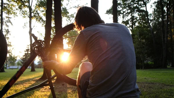 L'uomo gira e ripara una ruota su una bicicletta capovolta, il ragazzo ripara una bici al tramonto nel parco — Foto Stock