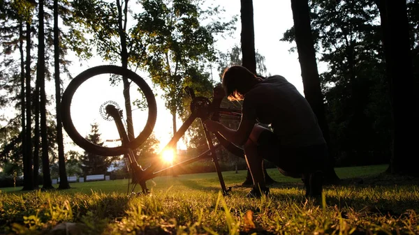 L'uomo gira la ruota e controlla la catena su una bici capovolta al tramonto nel parco — Foto Stock