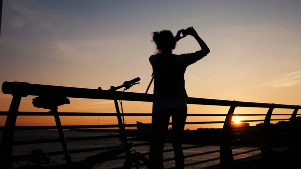 La silhouette di una ragazza fotografa il tramonto sul ponte d'estate verso il mare — Foto Stock