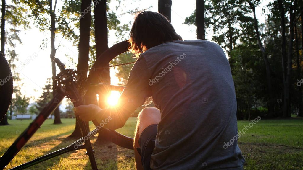 Man spins and repairs a wheel on an upturned bicycle, the guy repairs a bike at sunset in the park