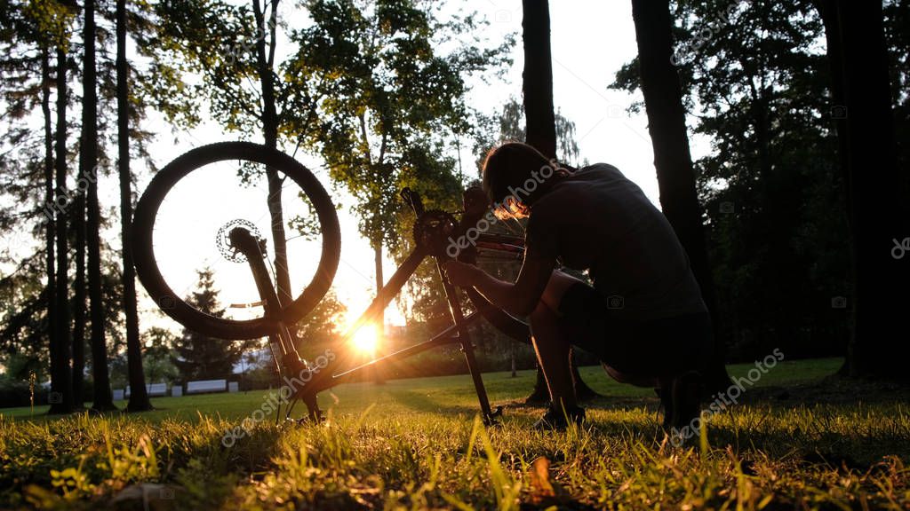 Man spins the wheel and checks the chain on an upturned bike at sunset in the park