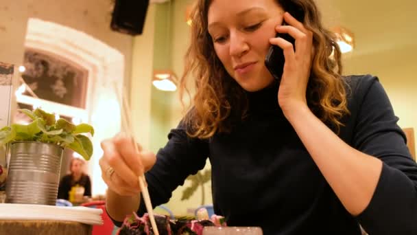 Attraente ragazza in un caffè vegetariano parlando al telefono e mangiando rotoli di cibo crudo — Video Stock