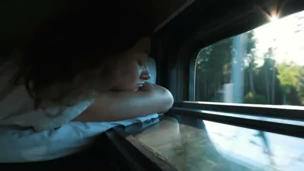Menina dorme em um trem com uma janela aberta ao pôr do sol, o vento sopra seu cabelo, os raios de sóis brilham pelas árvores — Vídeo de Stock