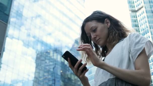 Дівчина використовує Інтернет на смартфоні, друкуючи текст на вулиці на тлі хмарочосів у бізнес-центрі — стокове відео