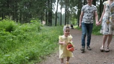 Çocuklu genç bir aile ormanda yürüyor. Küçük bir kız kovayla koni topluyor.