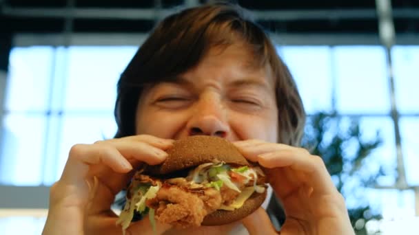 Preto olho cara come saboroso hambúrguer contra borrão janelas — Vídeo de Stock