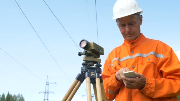 Topógrafo tipos en el teléfono inteligente y mira a través de theodolite — Vídeo de stock