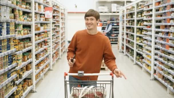 Chico bonito con un teléfono y una cesta de comida bailando en el supermercado — Vídeo de stock