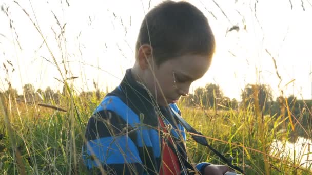 Poco boyconcentrated mira la pantalla de la cámara en la hierba al atardecer — Vídeo de stock