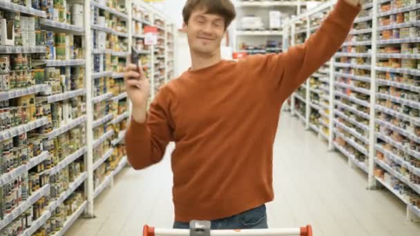 Счастливый парень в коричневой толстовке танцует держа смартфон в магазине — стоковое видео