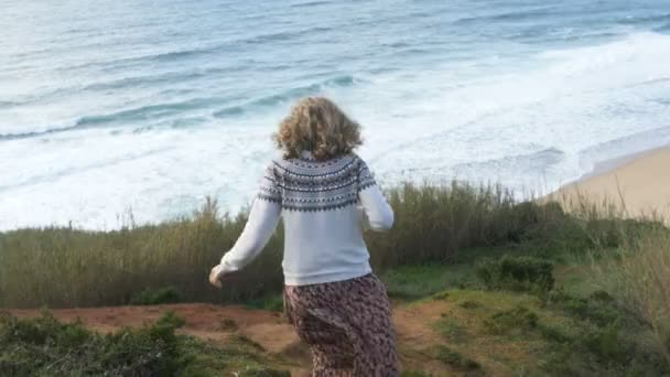 Девушка спускается вниз по холмистому пляжу к океану и фотографирует — стоковое видео
