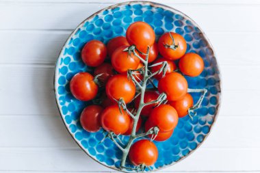 İştah açıcı olgunlaşmış domatesler, beyaz ahşap arka planda mavi bir kil tabakta bir dal üzerinde.