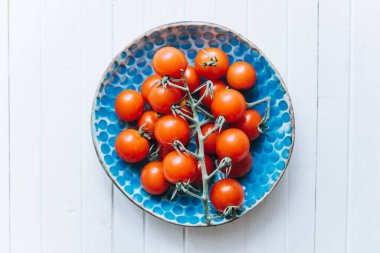 İştah açıcı olgunlaşmış domatesler, beyaz ahşap arka planda mavi bir kil tabakta bir dal üzerinde.