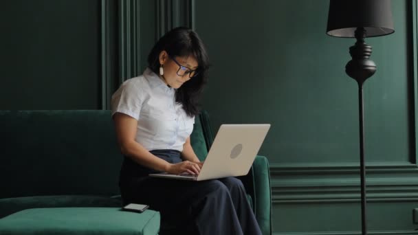 Wanita tersenyum dalam tipe blus pada laptop abu-abu duduk di sofa — Stok Video