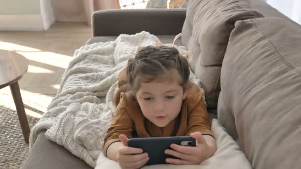 Маленька дівчинка інтенсивно дивиться мультфільми на свій мобільний телефон, лежачи на дивані — стокове відео